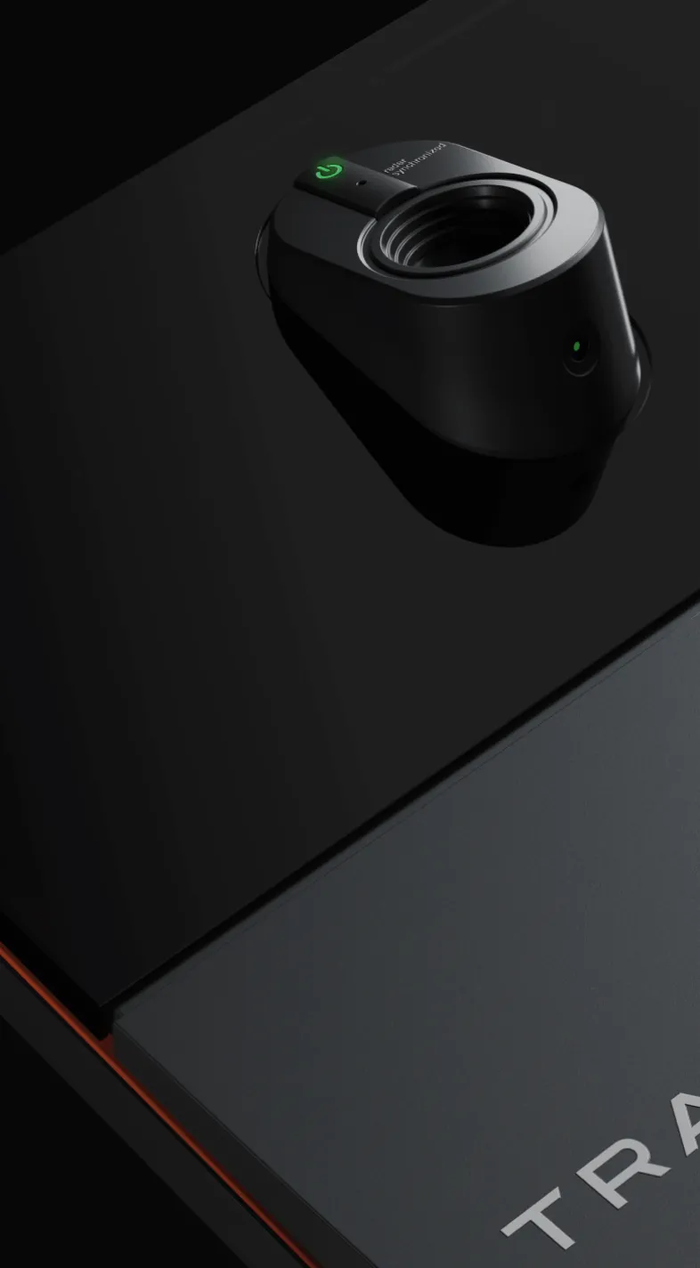 Close-up of a sleek black device with a lens and green indicator light on a dark surface, featuring the partial word "TRA" in white.