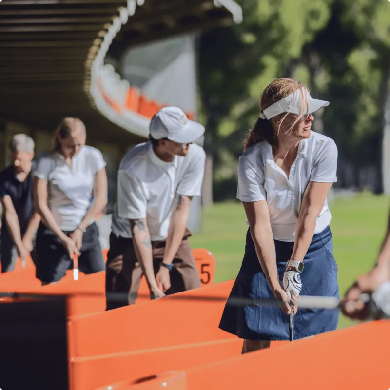 People in casual attire practicing golf swings at a driving range, with vibrant orange dividers and greenery in the background.